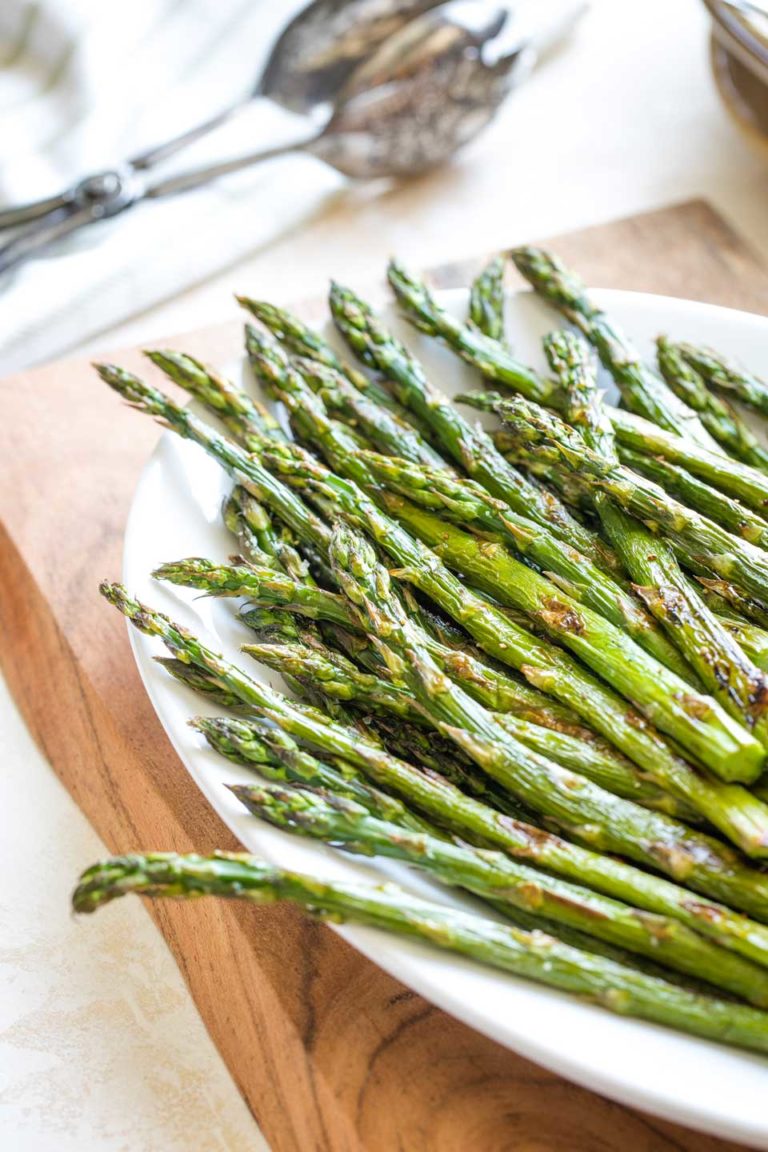Partial view of the top portion of the roasted asparagus spears after they're out of the oven and pile on a white serving platter that's sitting on a wooden cutting board.