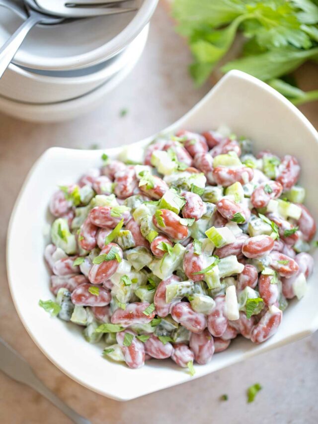 Overhead of a curved whi serving bowl with cut-out sides, filled with this bean salad recipe, with a stack of 3 little bowls and 4 forks in the background and a serving spoon alongside.