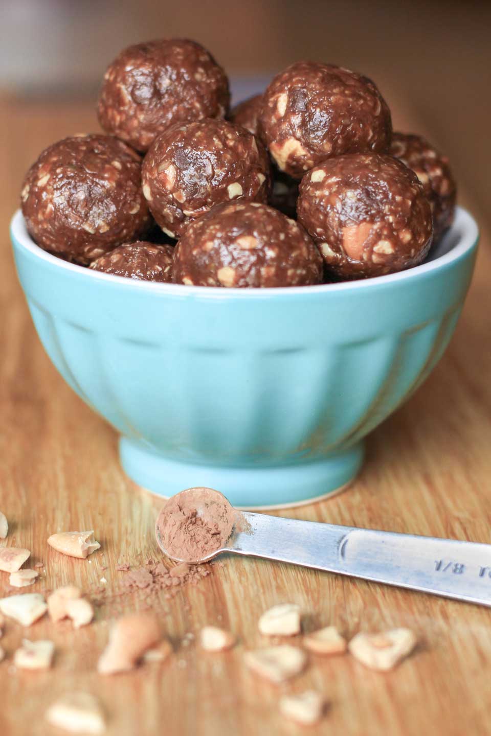 Aqua bowl full of peanut butter energy balls with crushed nuts and measuring spoon of chocolate in the foreground.