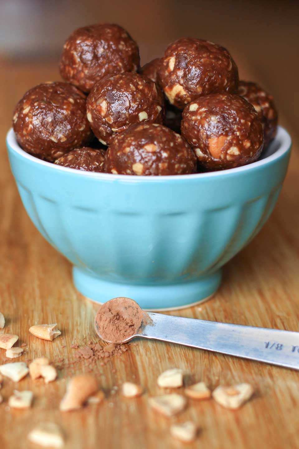 A batch of balls in a turquoise bowl with measuring spoon of cocoa powder and chopped peanuts in front of it.