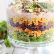 Side view of a clear trifle bowl with layers of salad, corn, tomatoes, cheese, and other Mexican ingredients.