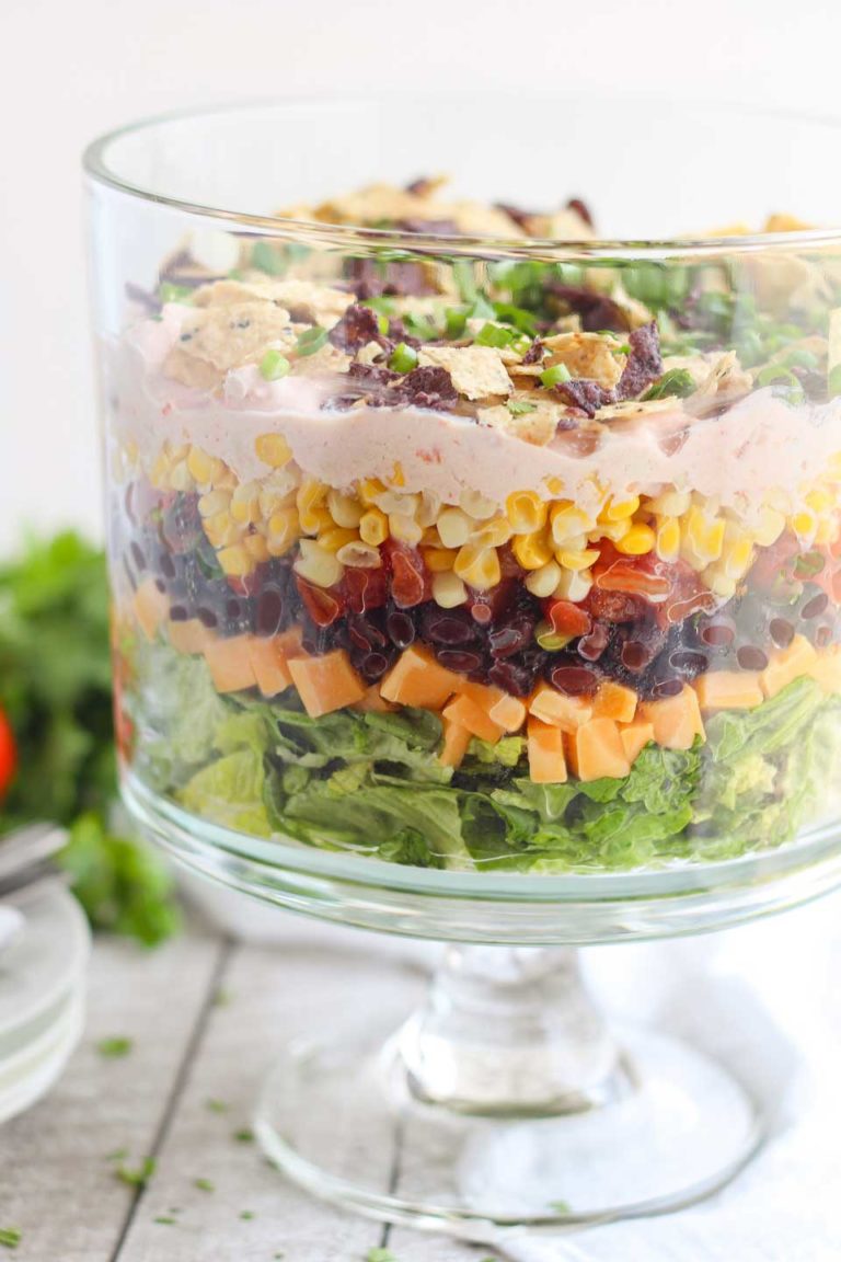 Side view of a clear trifle bowl with layers of salad, corn, tomatoes, cheese, and other Mexican ingredients.