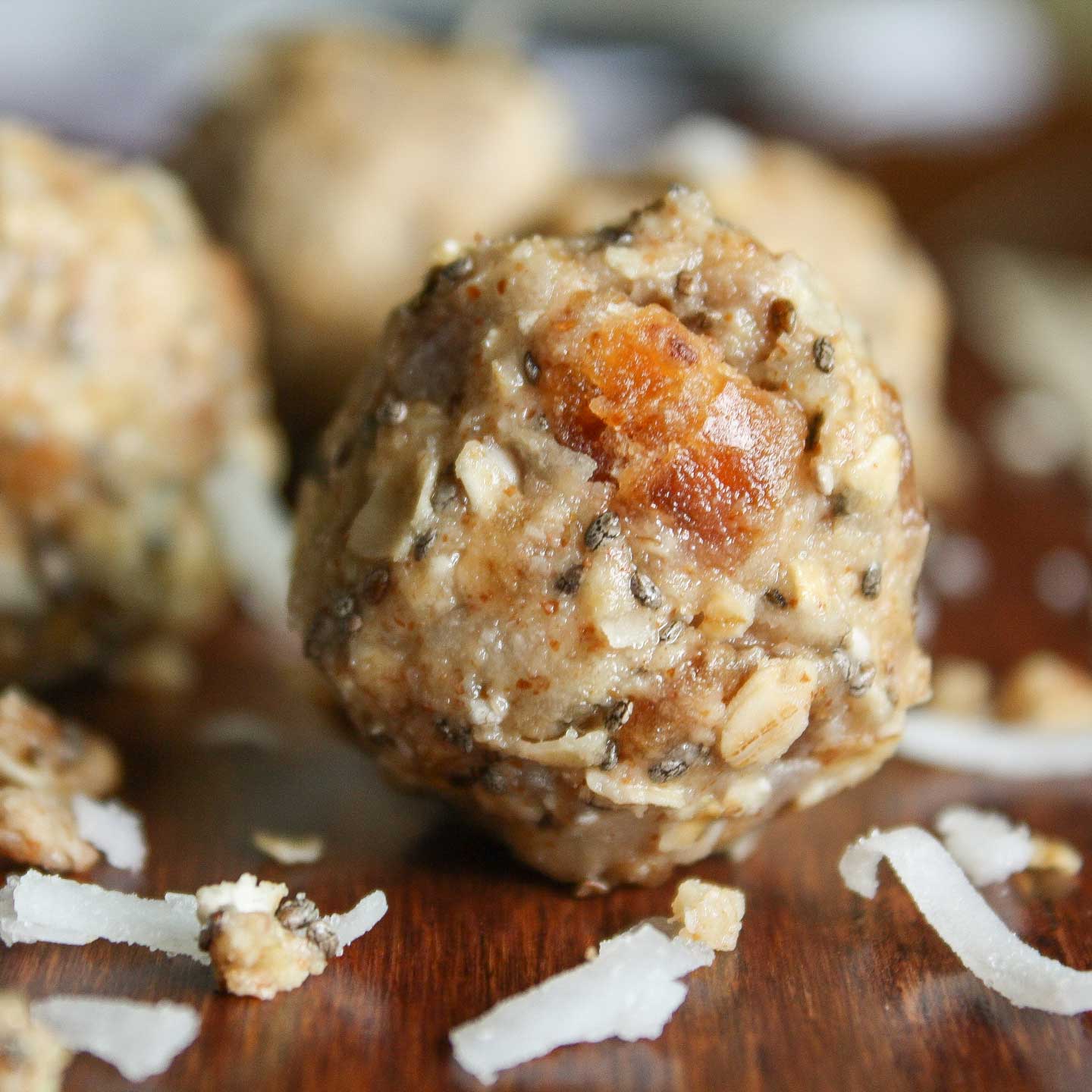 Closeup of finished energy bite, with several more balls in background and ingredients like coconut flakes laying around.