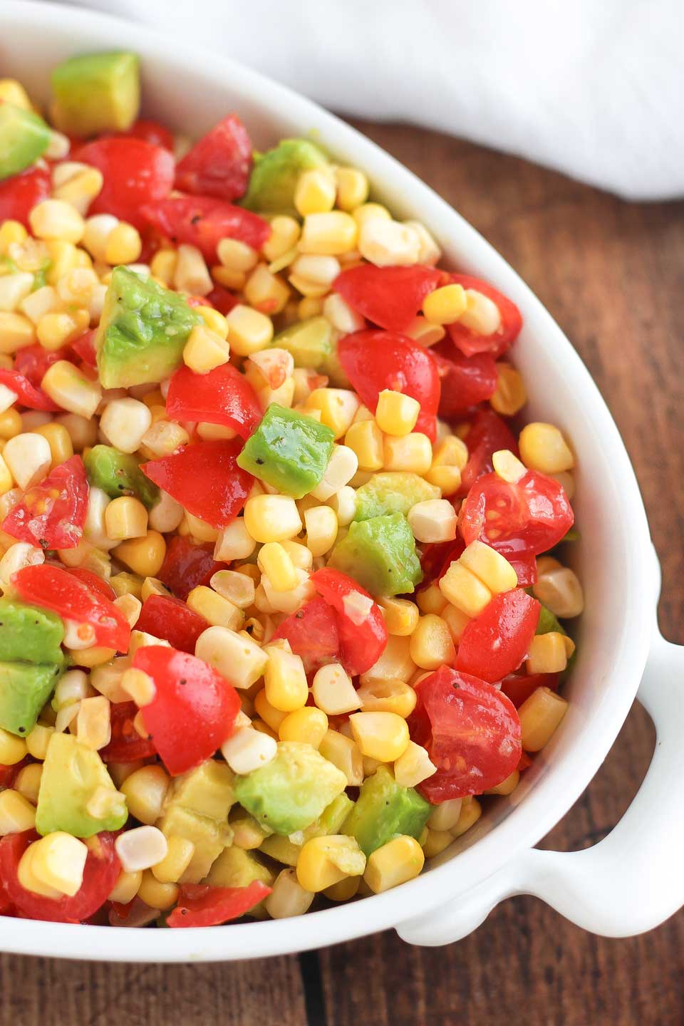 Overhead of the right half of an oval bowl filled with fresh corn salad with chunks of tomato and avocado.