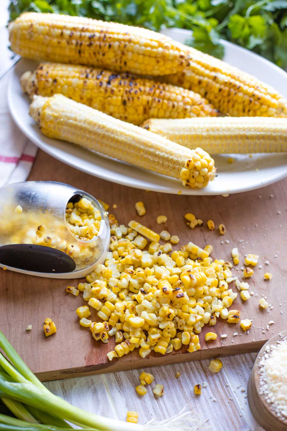 A pile of kernels cut off nearby corn cobs laying on a platter, with a corn stripper and other corn salad ingredients.