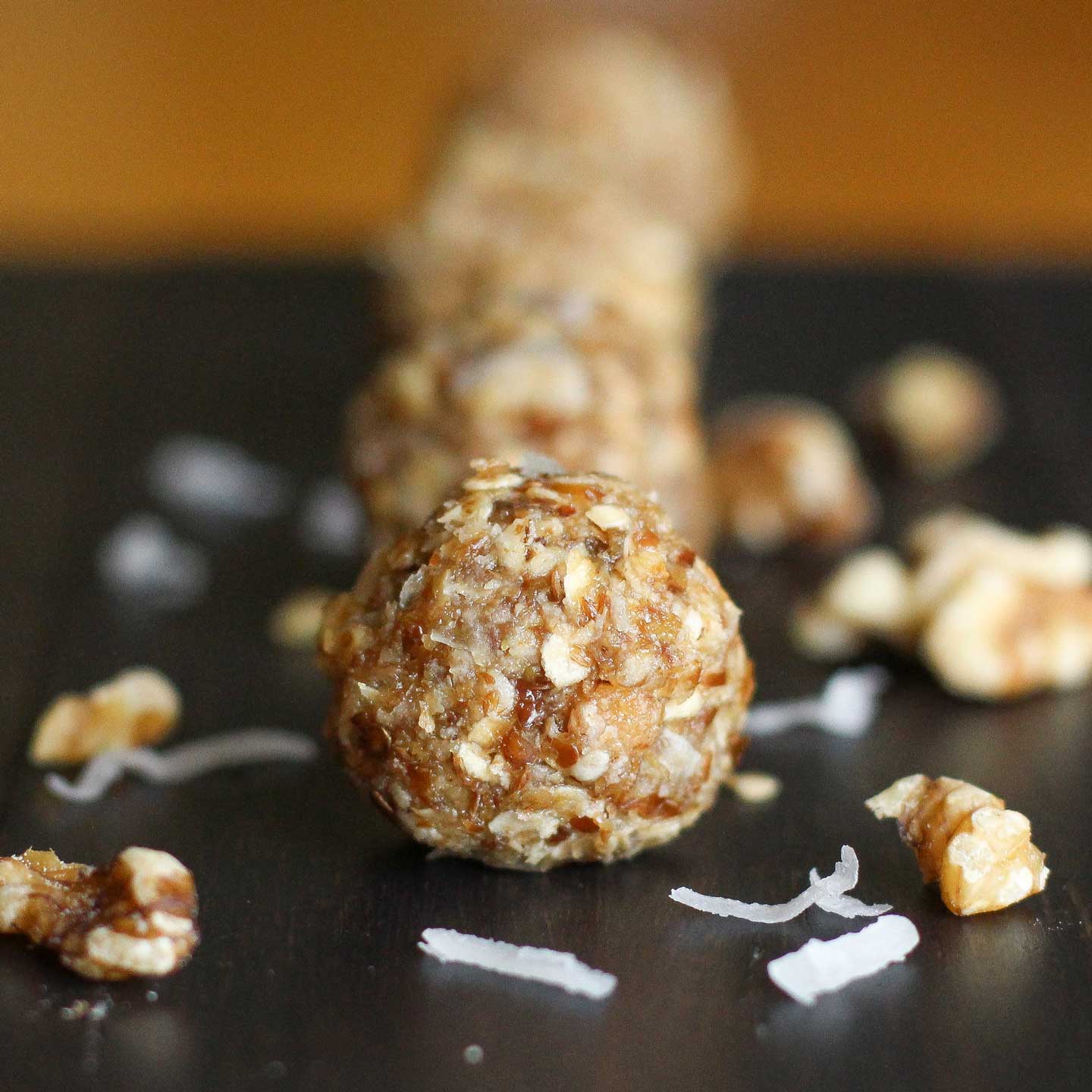 Closeup of the first ball in a row of 4 energy bites, laying on black wood with extra nuts and coconut sprinkled around.