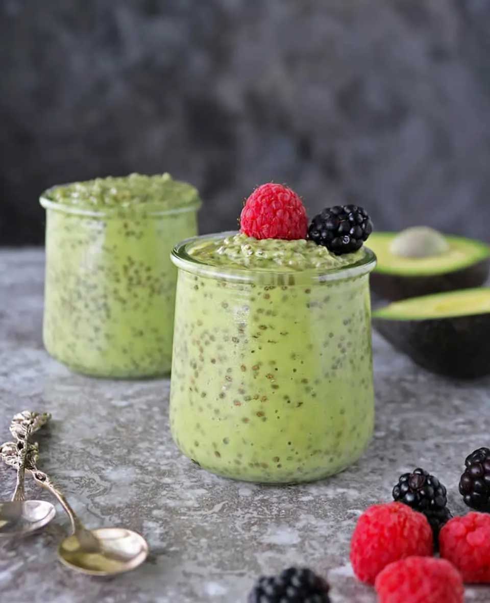 Side view of two filled jars garnished with berries, with an open avocado, two spoons and more berries nearby.