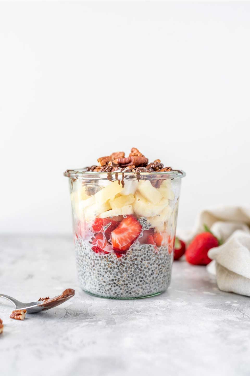 Side view in a glass jar so you can see the bottom layer of chia pudding, topped with sliced strawberries, then pineapple and bananas, then pecans and chocolate drizzle.