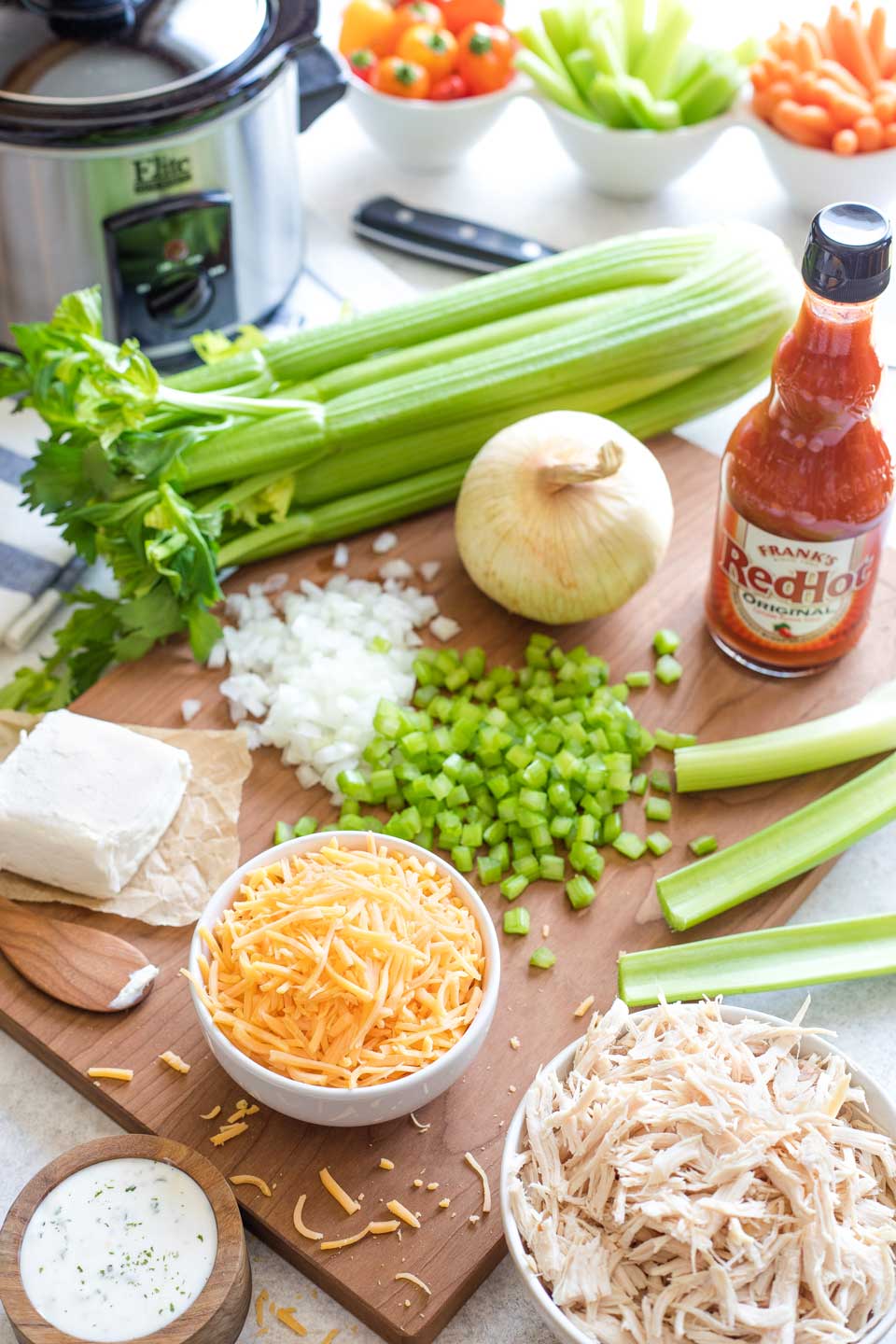 Ingredients like Frank's Red Hot Sauce and chopped vegetables on a wooden cutting board near empty crockpot.