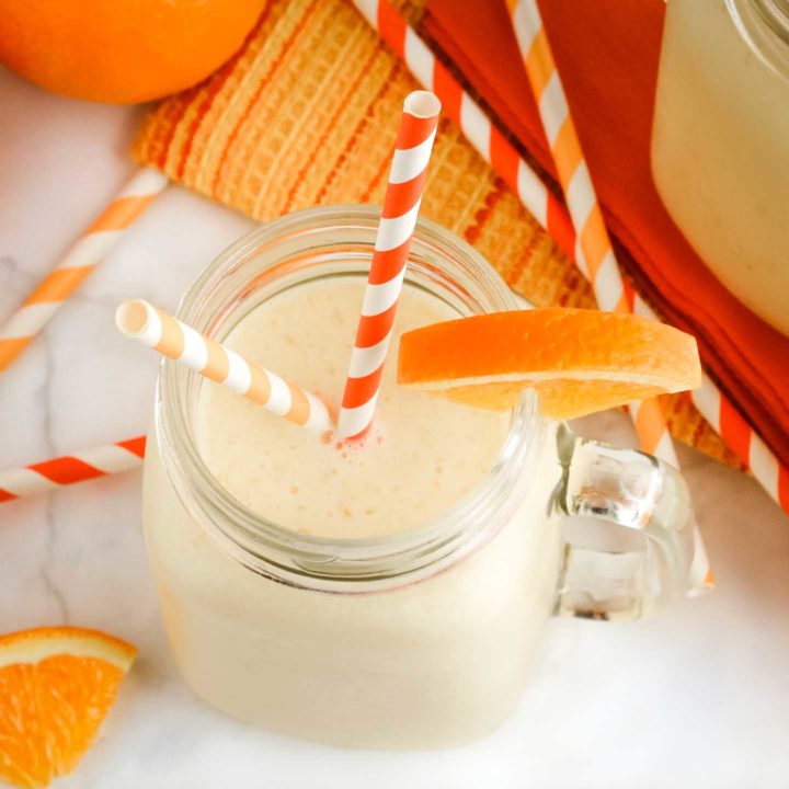 Finished smoothie in a clear glass mug with two striped orange straws and a wedge of orange on the rim.