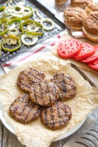 Five grilled hamburgers stacked on a plate with buns and Italian veggie toppings nearby.