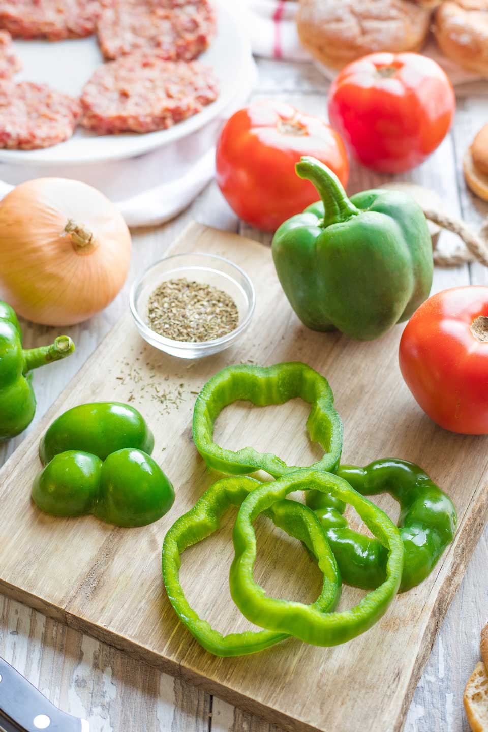 Ingredients: green pepper rings on cutting board, surrounded by tomatoes and Italian spices with buns and raw burgers in background.