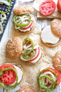 Overhead of 5 Italian hamburgers in various stages of being piled with grilled veggies, toppings and buns.