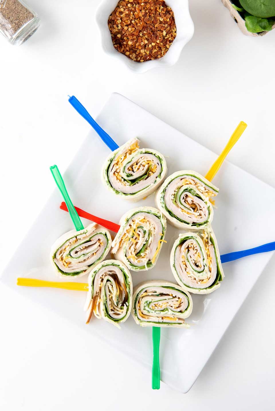 7 tortilla roll-ups on a square white plate, skewered with brightly colored appetizer picks.