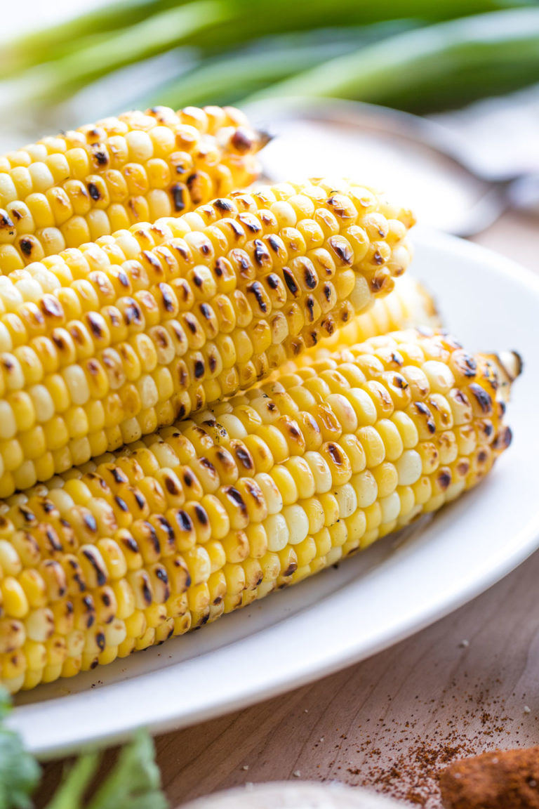 5 ears of grilled corn on the cob on a white platter, with pretty greens and spices nearby.