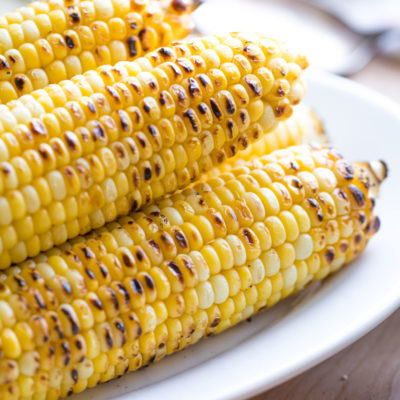 Serving platter of sweet corn that’s been grilled and has caramelization, with other dinner ingredients nearby.