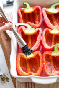 Hand brushing oil onto un-stuffed peppers in baking dish.