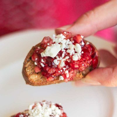 Fingers holding one crostini topped with cranberry dip and sprinkled with goat cheese with another on plate in background.