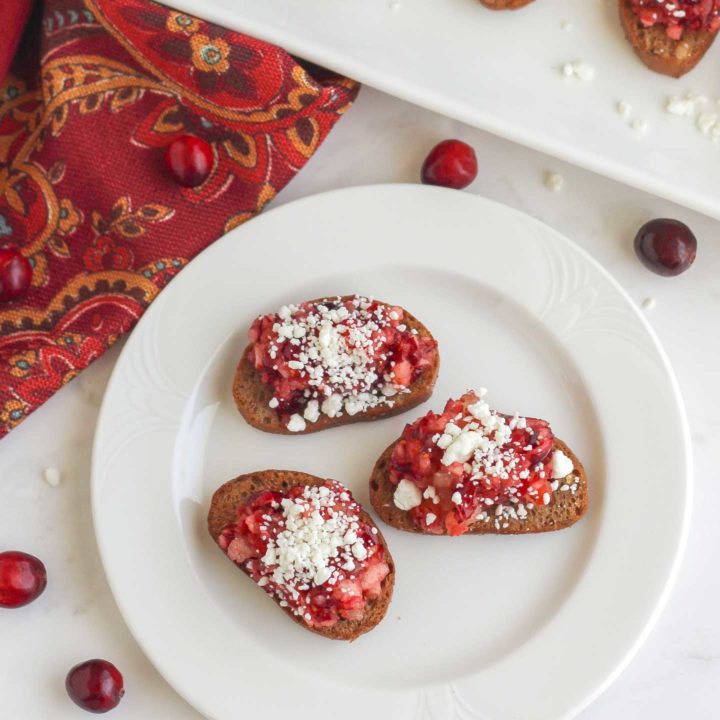 Flatlay of 3 cranberry appetizer toasts on a white plate with more on a serving tray in background.