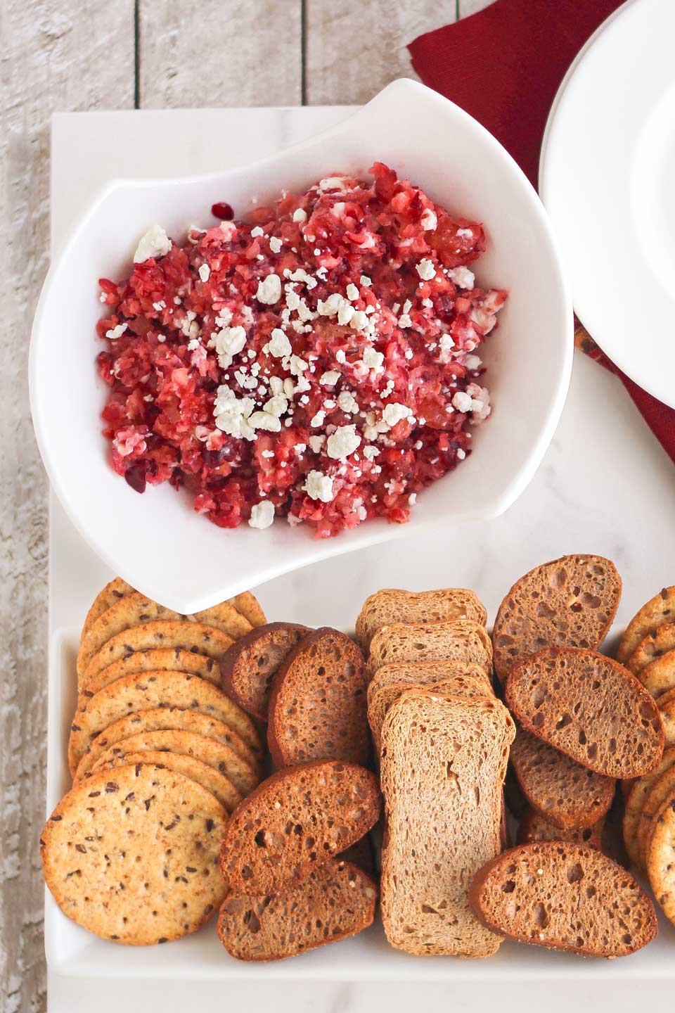 Cranberry dip presented in serving bowl, sprinkled with goat cheese, with crostini and crackers ready for dipping.