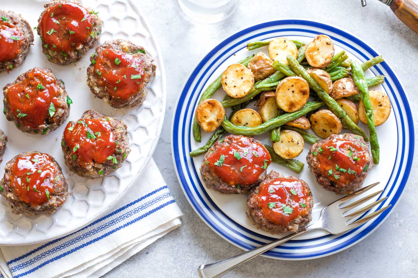 Three mini meatloaf patties served on blue-rimmed plate with vegetable, with a white serving platter full of more meatloaf alongside.