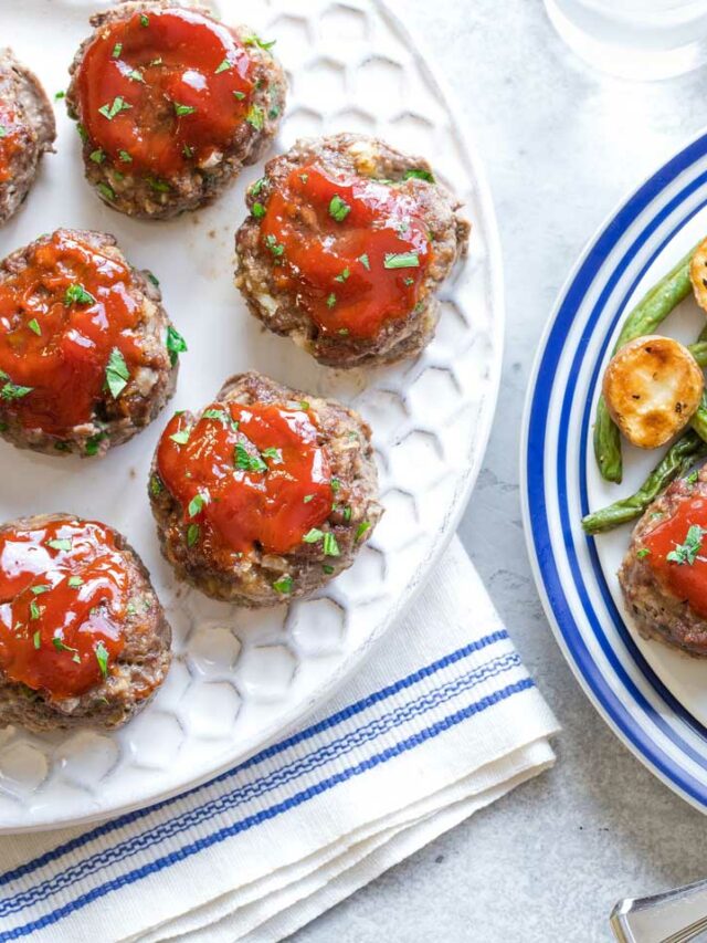 Three mini meatloaf patties served on blue-rimmed plate with vegetable, with a white serving platter full of more meatloaf alongside.