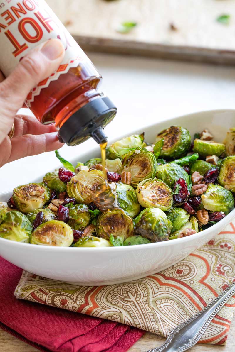 Hand squeezing bottle of hot honey to drizzle honey over roasted Brussel sprouts in serving dish.