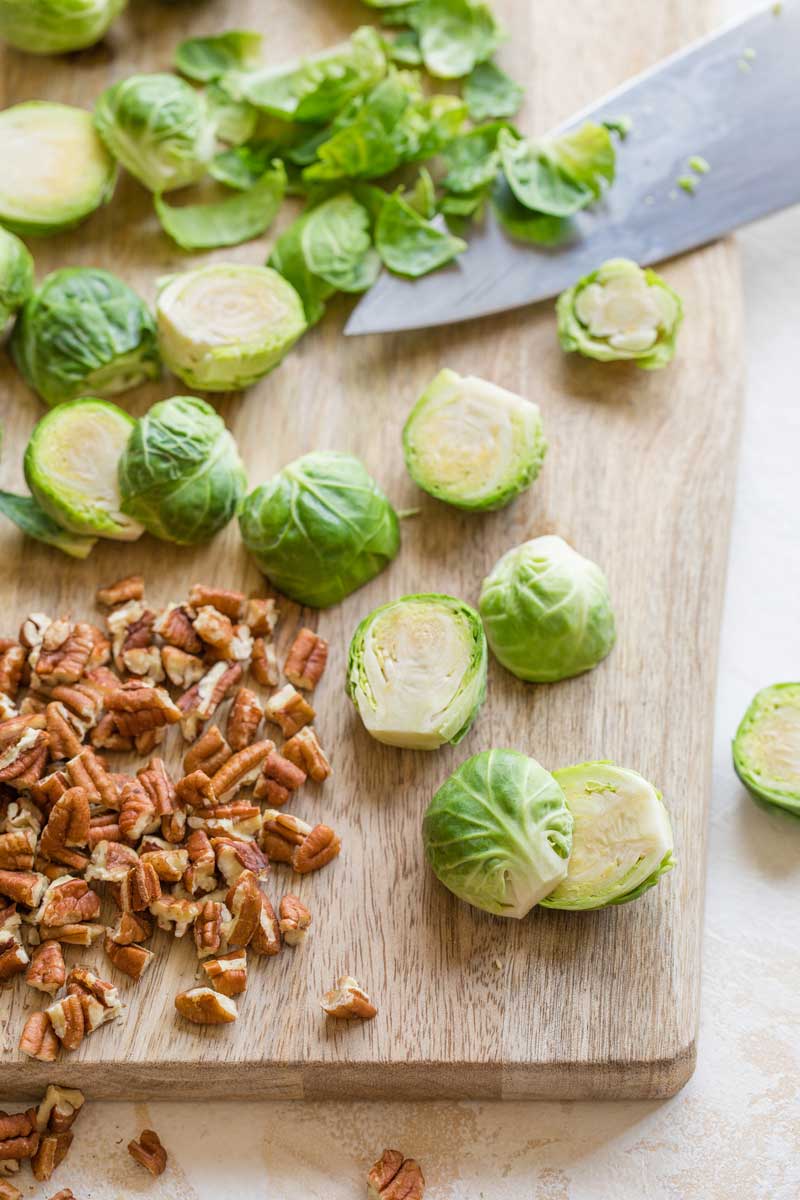 Showing how to trim ends and cut Brussels sprouts in half vertically, on cutting board alongside pecan pieces.