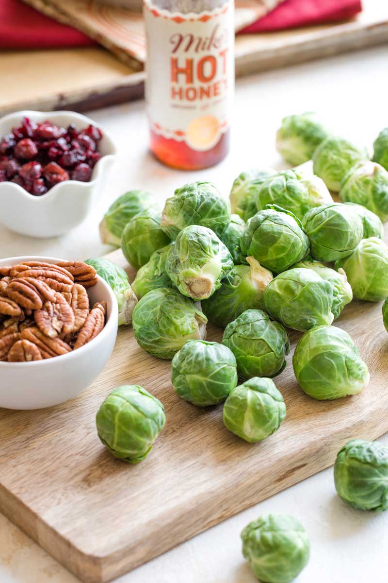 Raw, uncut Brussels sprouts cascading across cutting board with bowls of pecans and cranberries and a bottle of hot honey nearby.