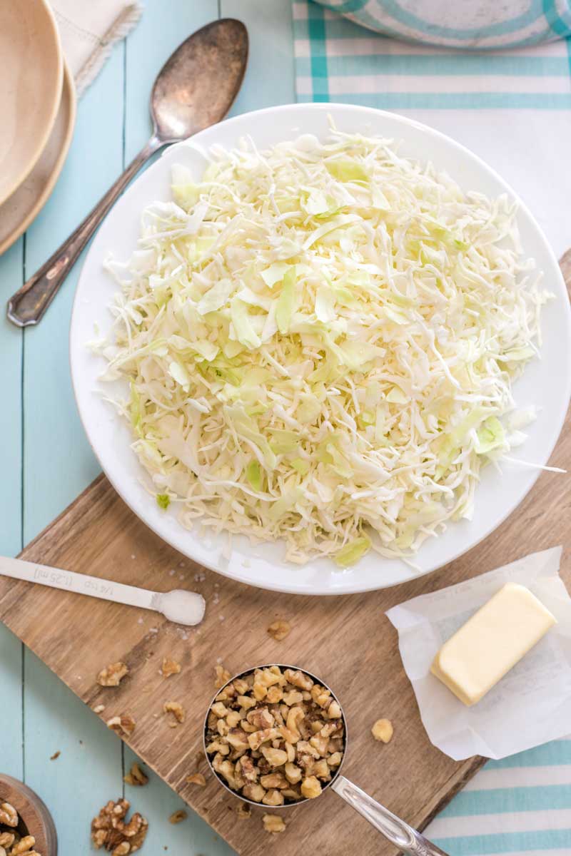 Overhead of shredded, raw cabbage, stick butter, chopped nuts and salt on cutting board, ready for cooking.