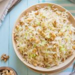 Overhead of serving bowl full of cooked cabbage on light blue background, with serving items and extra nuts near.