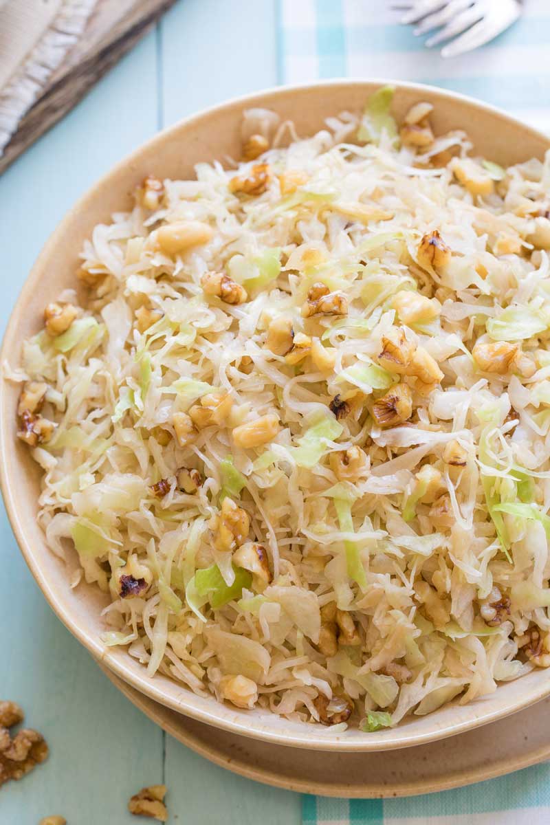 Overhead closeup of tan serving bowl of sauteed cabbage on light blue background and cloth, garnished with extra nuts.