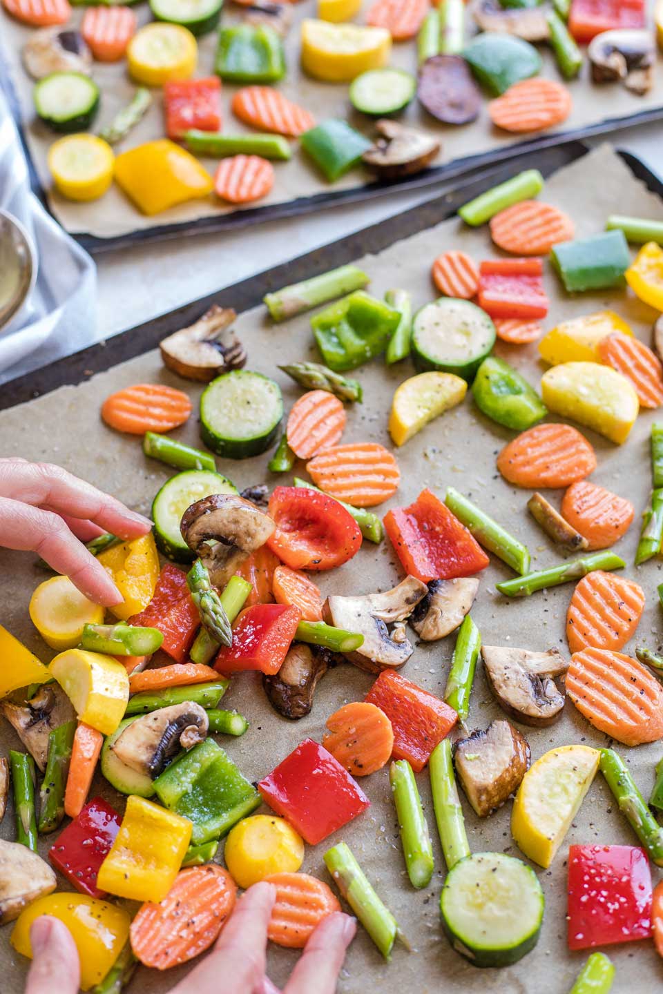 Two hands tossing vegetables with oil and seasonings on a brown parchment-lined sheet pan before going in oven.