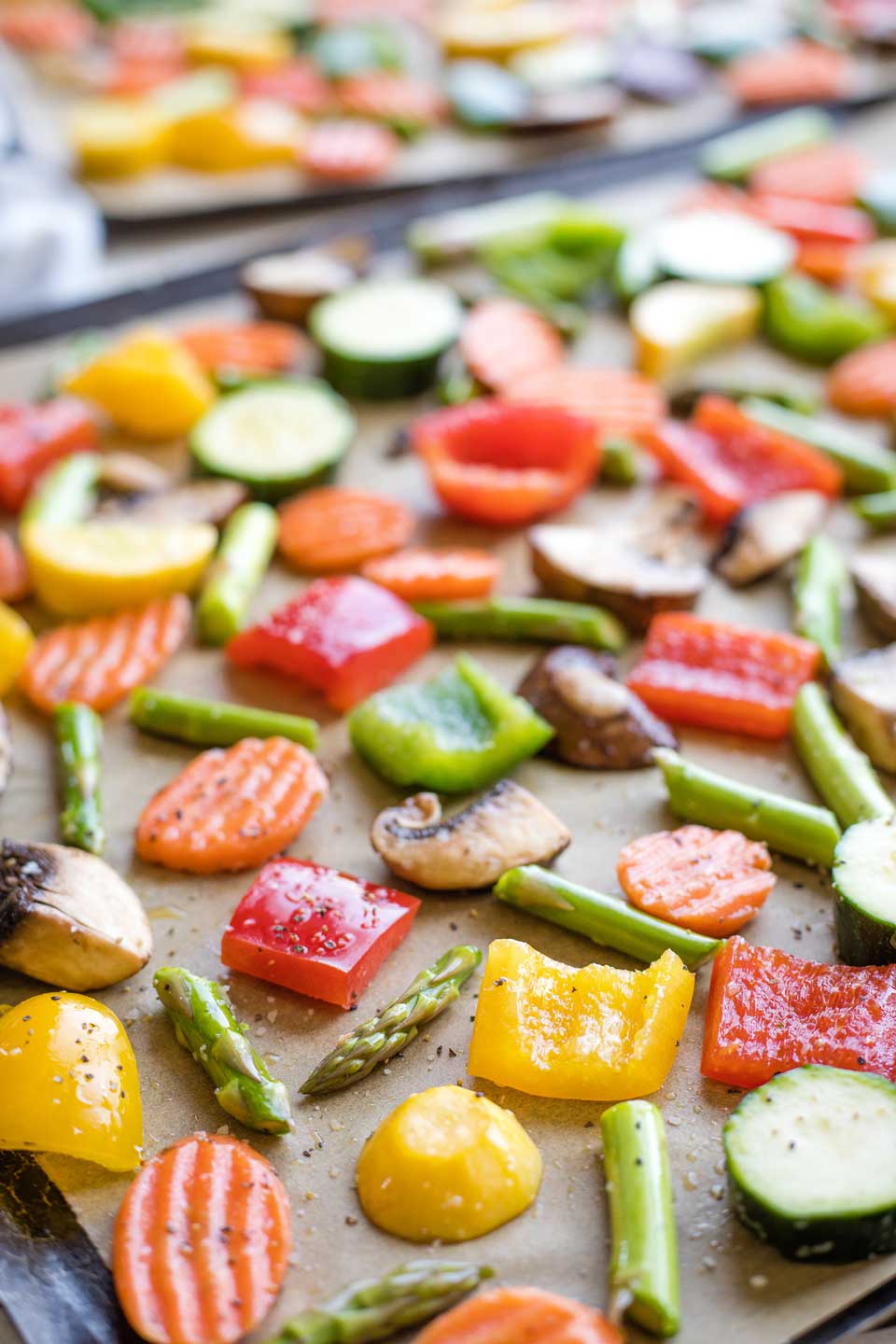 Raw vegetables coated in oil and seasonings, spread out on two sheet pans before being roasted.