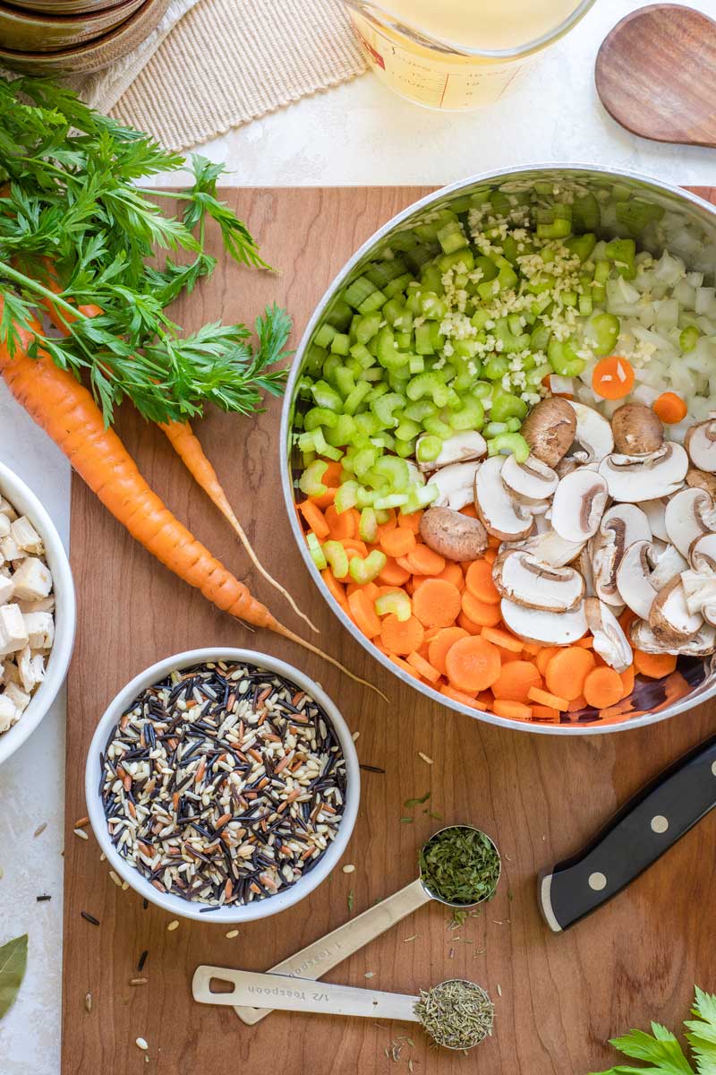 Overhead of soup pot full of raw veggies on cutting board with bowl of rice, spoonfuls of herbs and whole carrots.