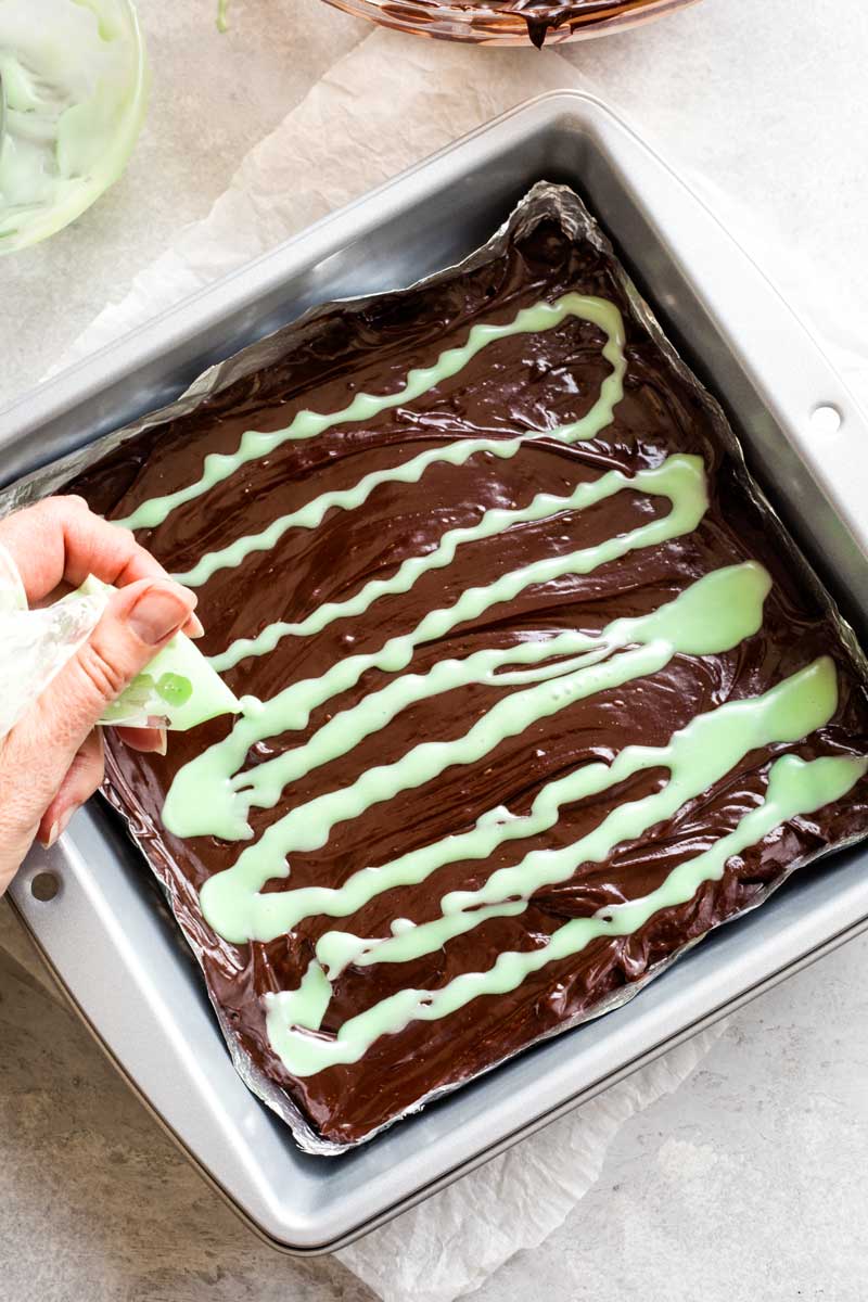 Hand squeezing baggie of mint-colored drizzle onto pan of warm chocolate in a zigzag pattern.