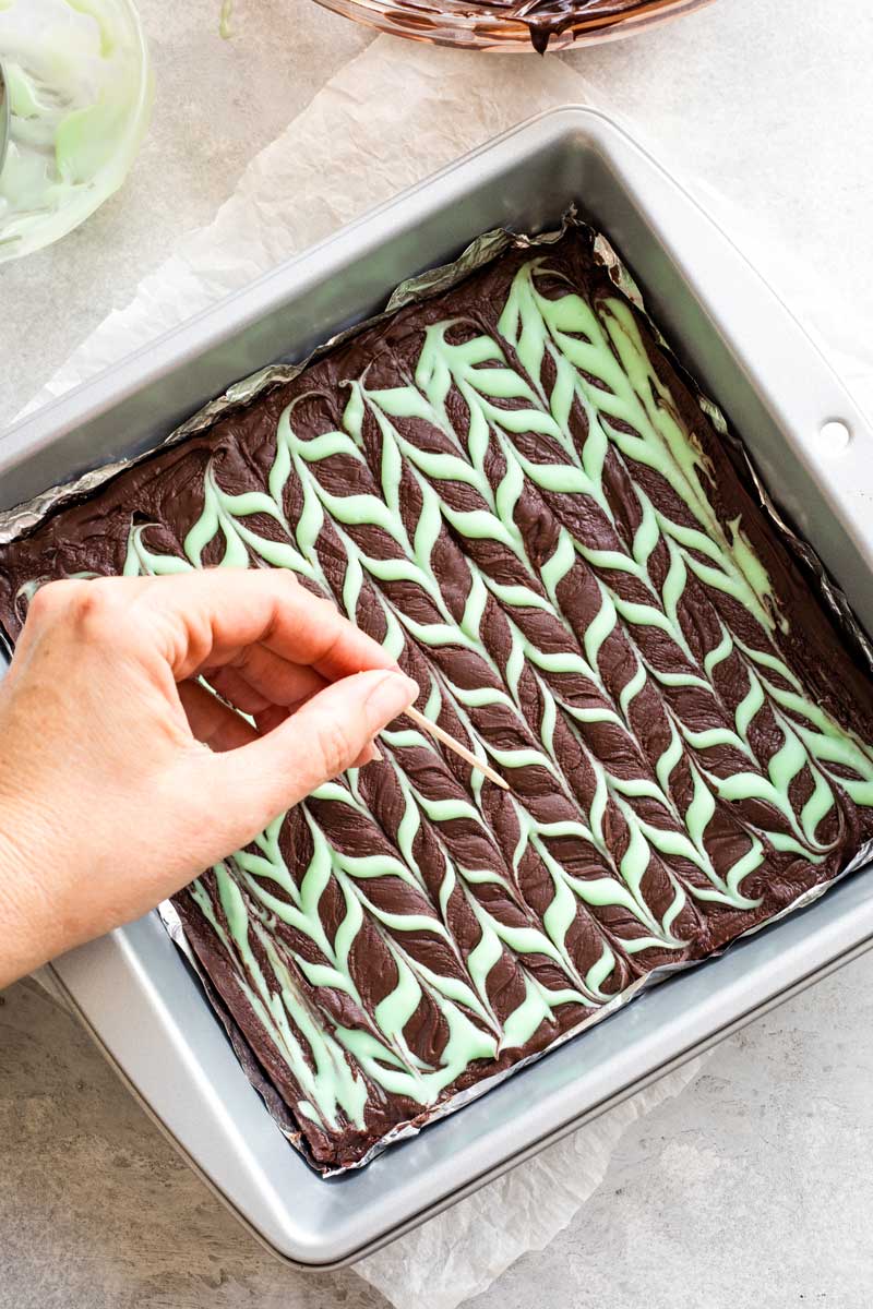 Hand dragging toothpick through mint colored drizzle to make design in fudge.