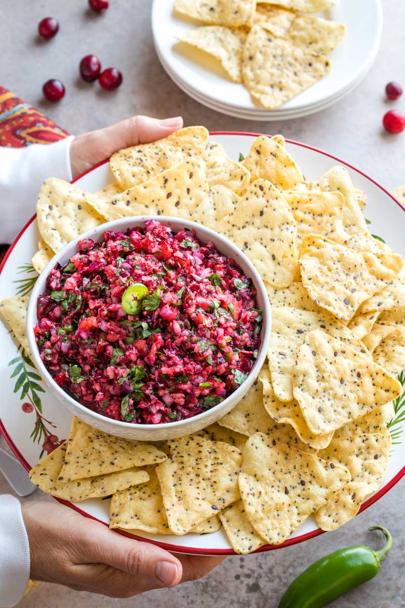Two hands holding holiday party platter of tortilla chips and bowl of salsa, with extra jalapeno, cranberry and plate of chips visible below.
