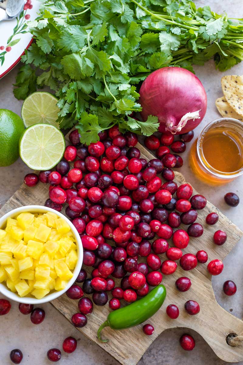Overhead of ingredients for the salsa: a cutting board with a cascade of cranberry, a bowl of chopped pineapple, limes, jalapeno, honey and cilantro.,