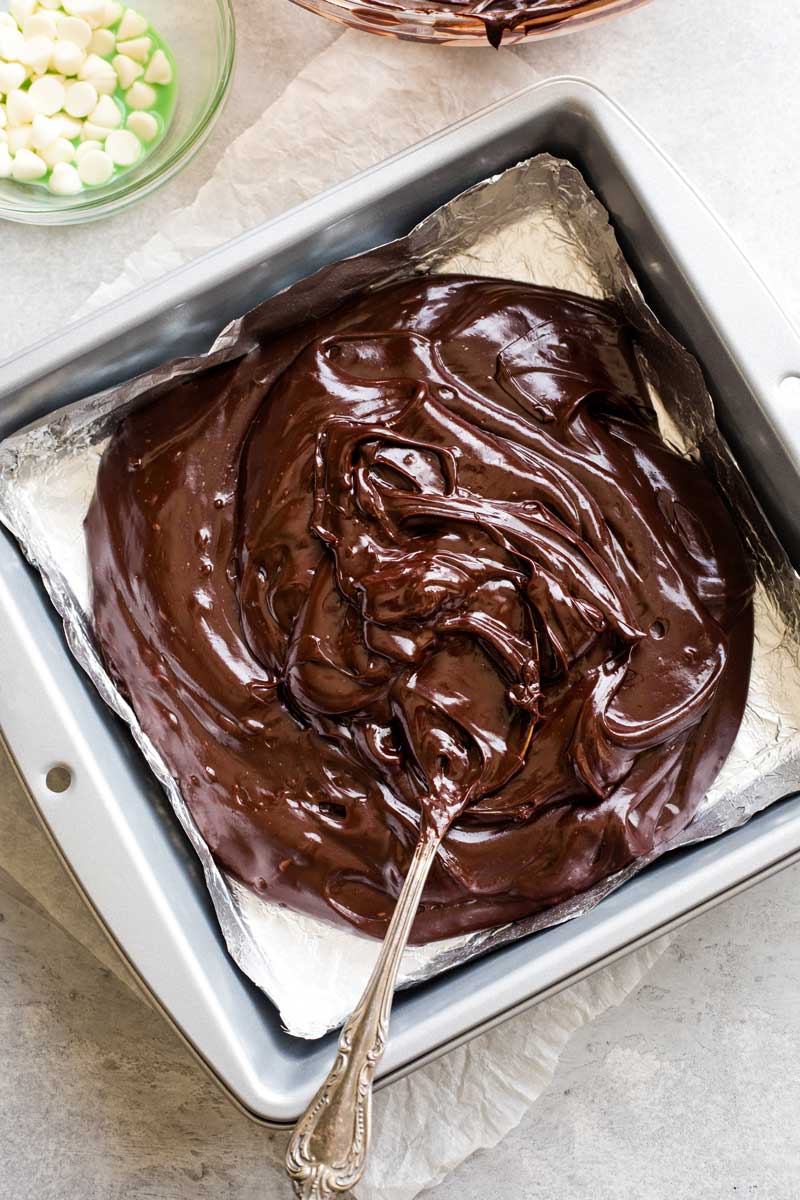 Mint chocolate mixture poured into foil-lined fudge pan with a spoon beginning to smooth it out.
