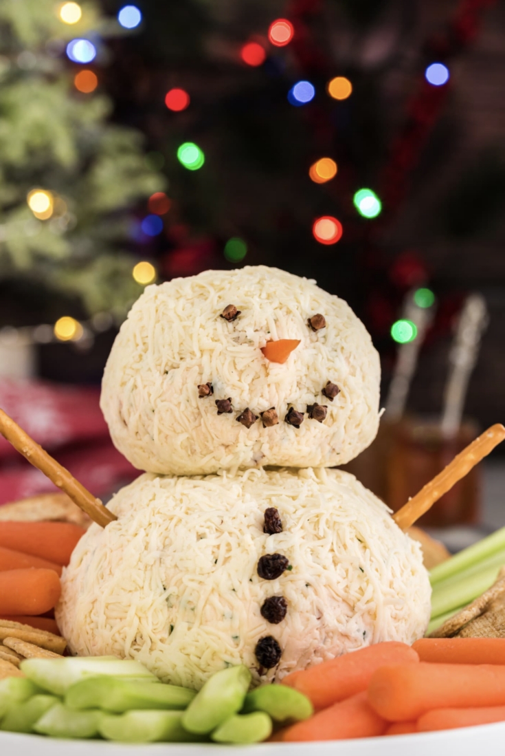Snowman in center of platter of crackers and crudites with twinkling Christmas lights in the background.