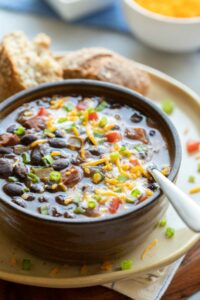 Black Bean Soup served in brown bowl on yellow plate, with spoon dipped in and roll on the side.