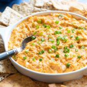 Healthy Buffalo Chicken Dip recipe presented for a party, with spoon in, chips all around, and vegetable strips for dipping in the background.