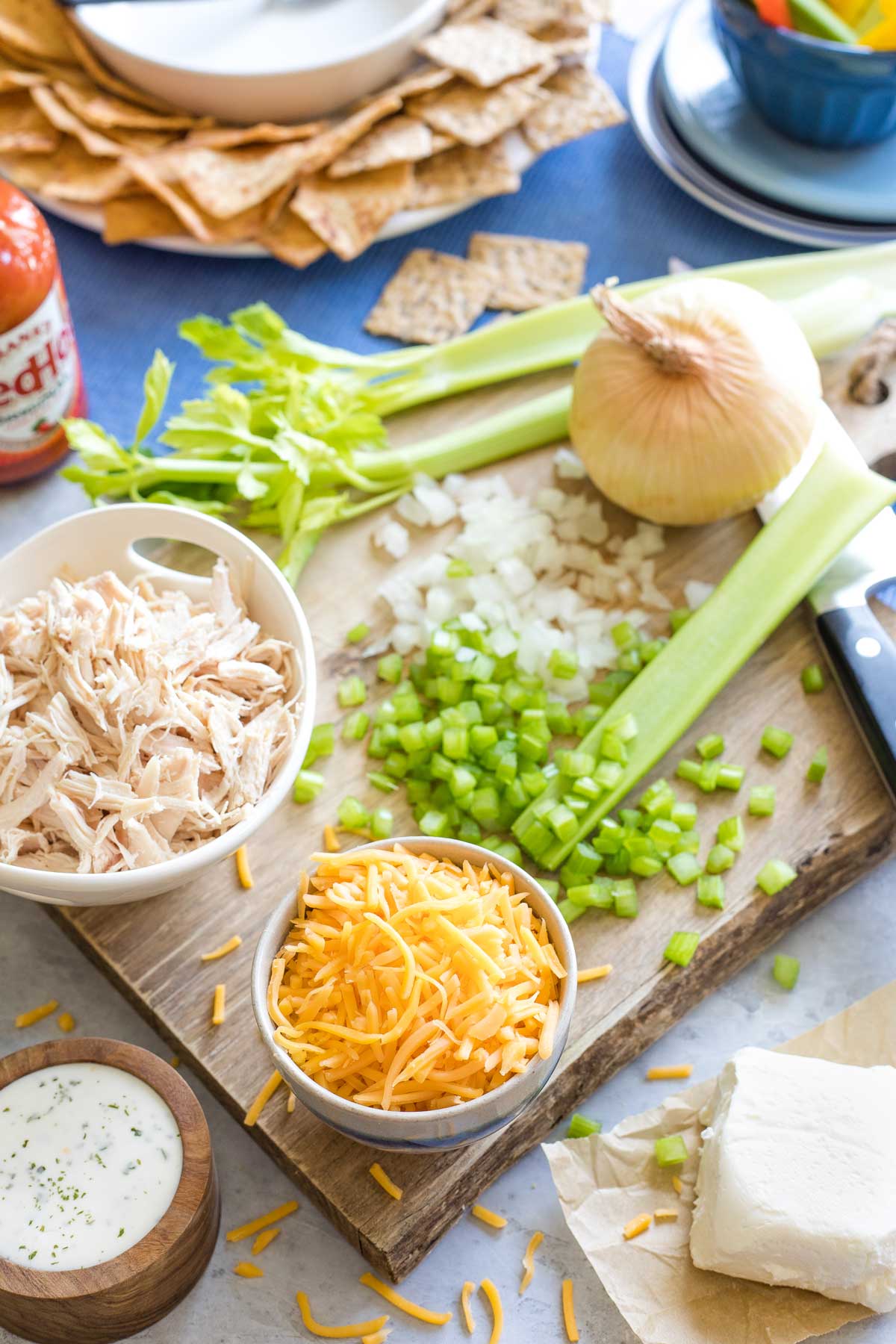 Ingredients on a wooden cutting board: shredded cheese and chicken, veggies, ranch and cream cheese, with a bottle of buffalo hot sauce in the background with dippers.