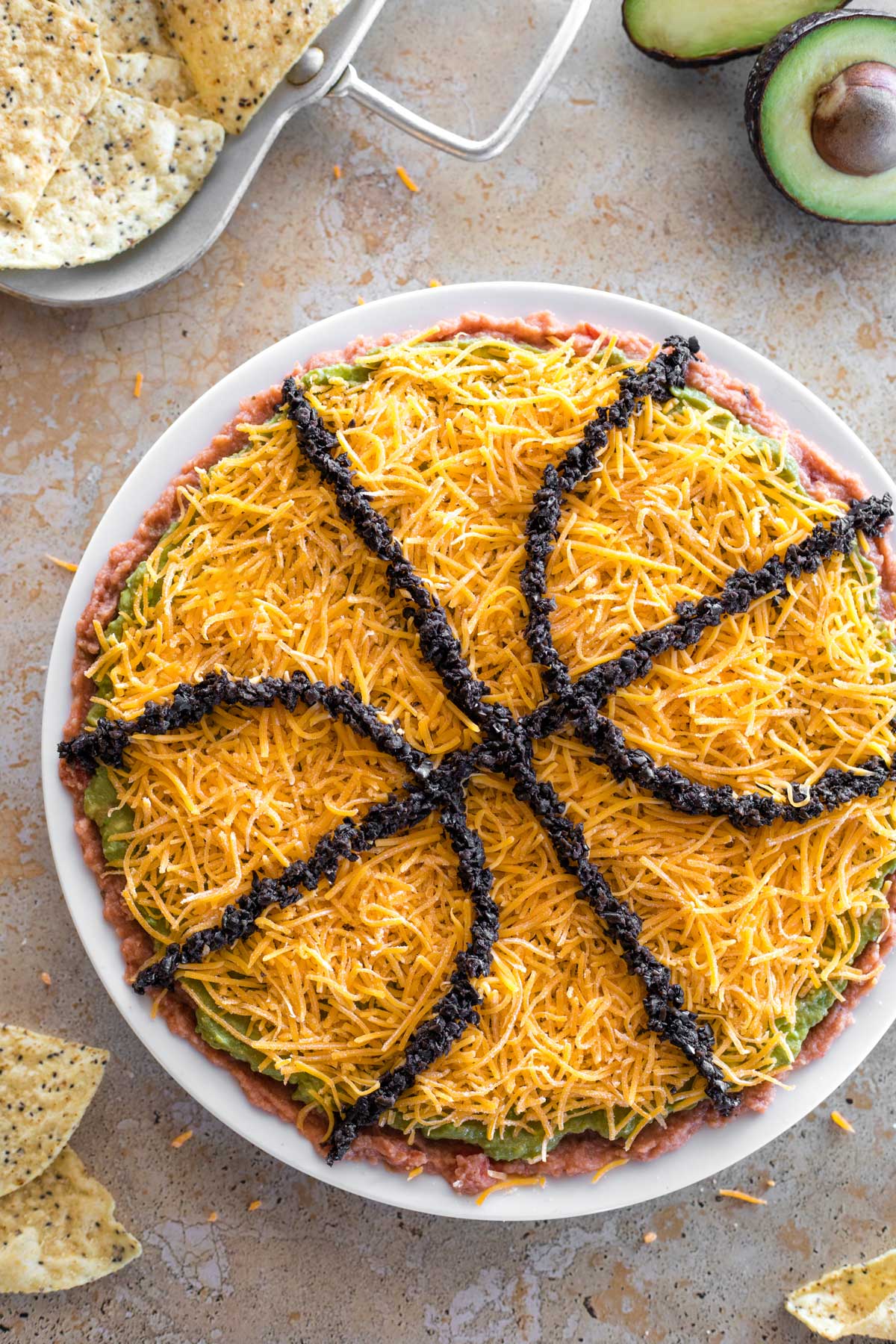 Overhead of finished March Madness layered dip, with tortilla chips and avocado halves decoratively in background.