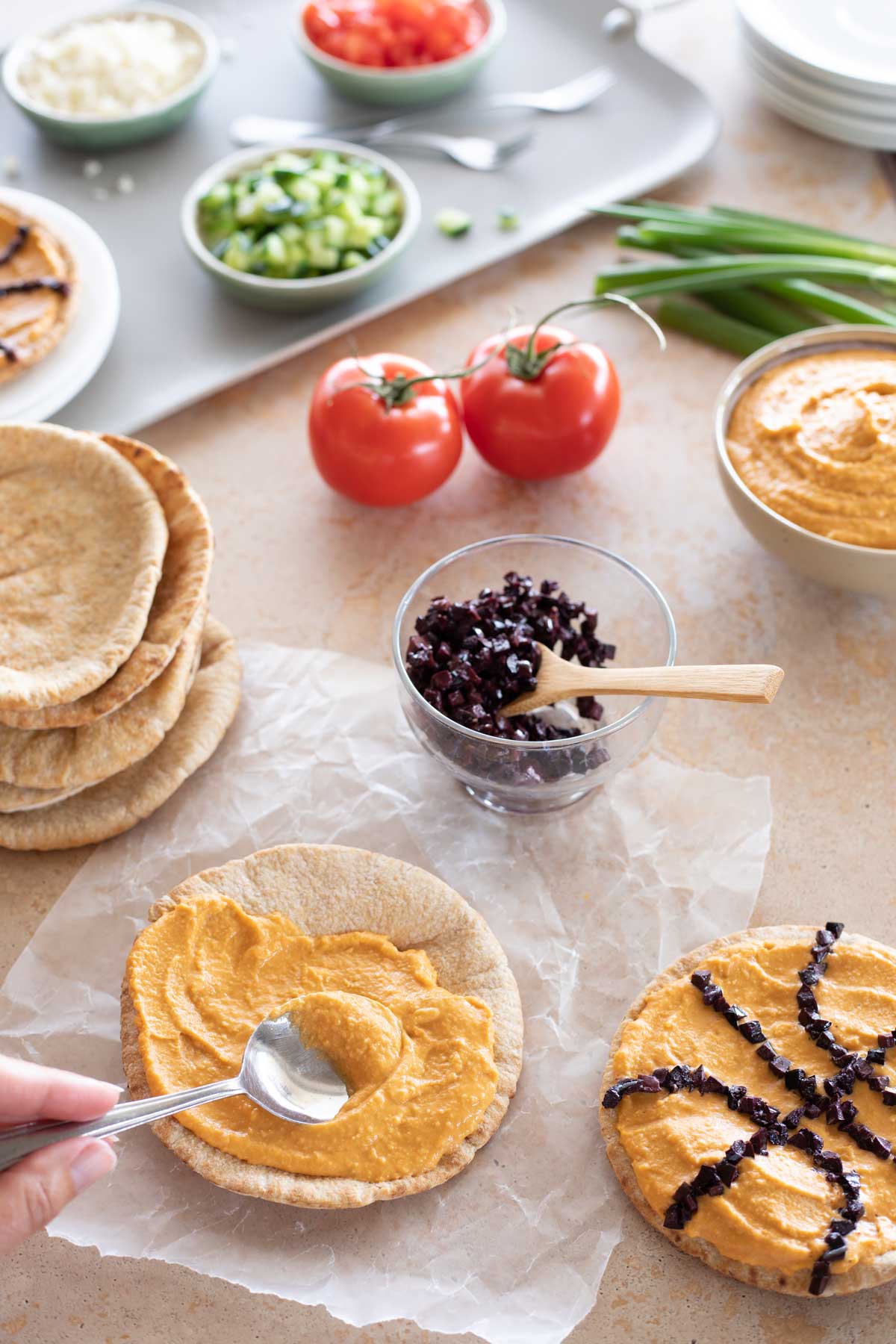 Fingers using spoon to spread hummus on pita, with other ingredients nearby.