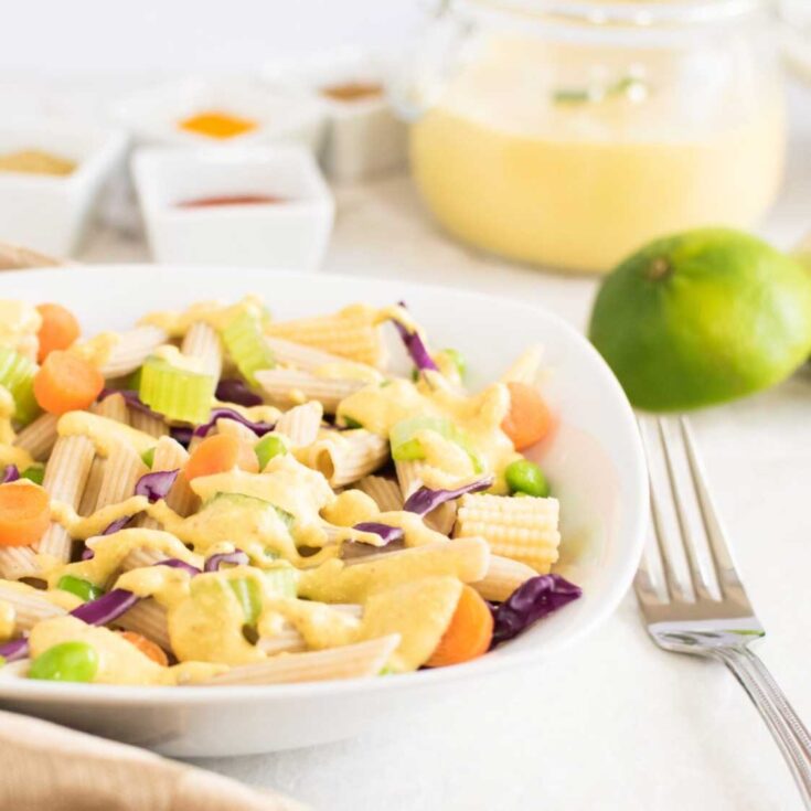 Closeup side view of right side of pasta bowl filled with salad, with fork alongside and extra ingredients in background.