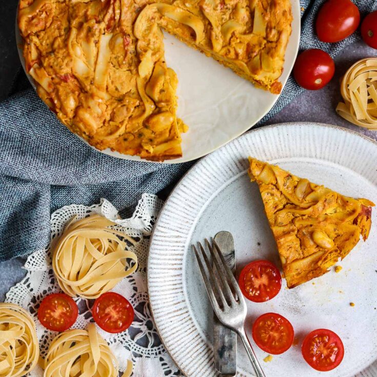 One wedge of frittata on a plate near the rest on a second plate with twirls of uncooked, dry vegan pasta and tomatoes nearby.