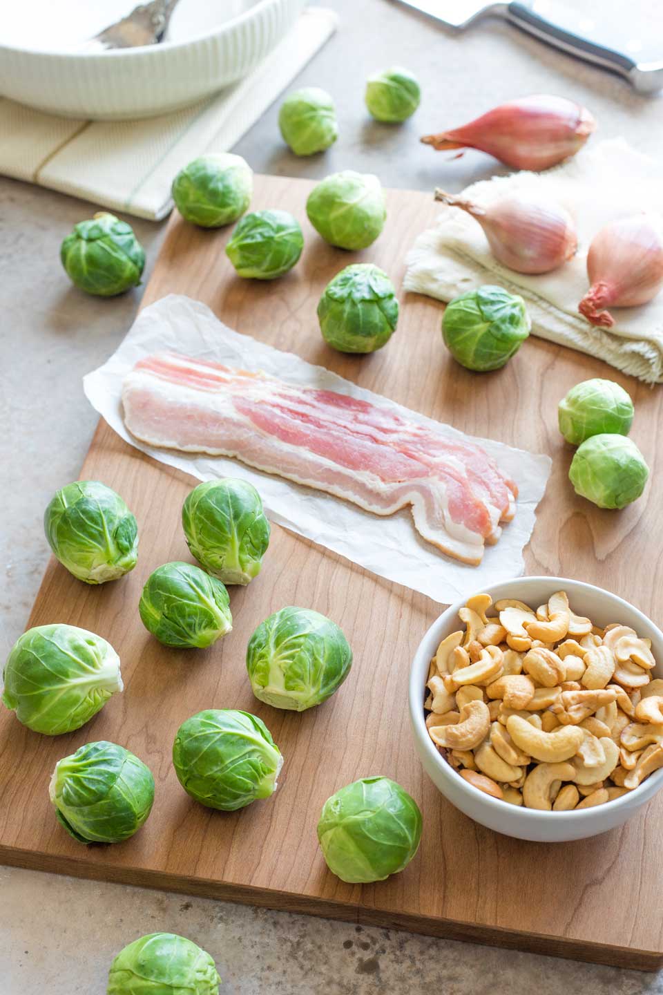 Whole Brussel sprouts scattered on cutting board with slices of bacon, unpeeled shallots and a bowl of cashews.