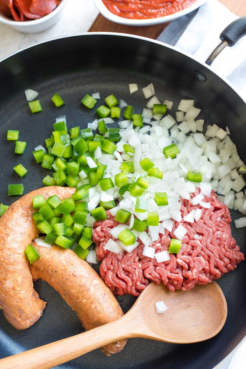 Raw onions, green pepper, ground beef, and Italian sausage links in large skillet with wooden spoon ready to begin cooking.